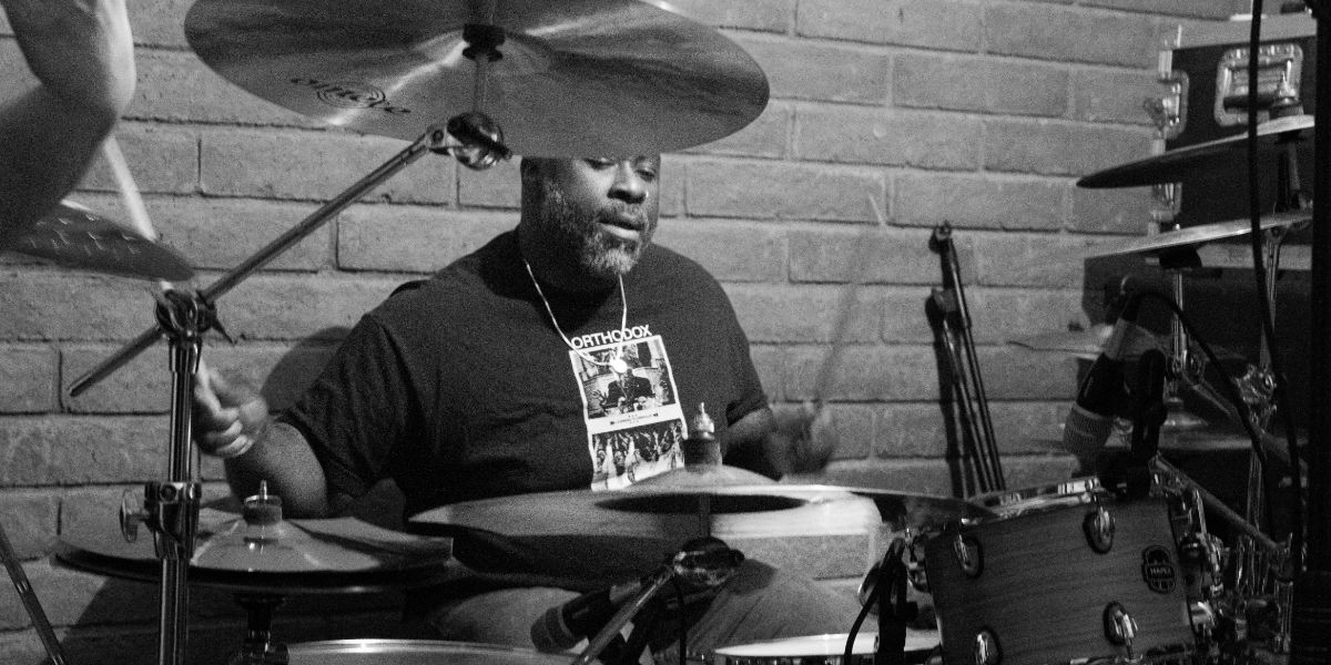Drummer actively playing full kit, cymbals prominent. Brick wall. GoatHead Audio drum recording.
